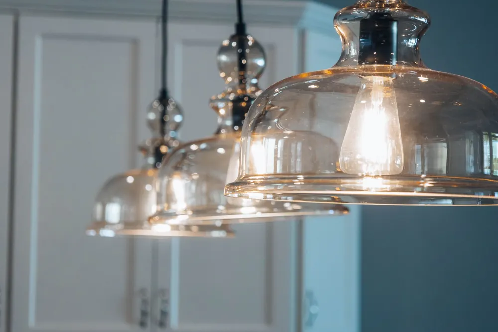 Glas-Pendelleuchten mit Fadenlampen hängen in einer Reihe vor weißen Küchenschränken, die vorderste leuchtet warm.