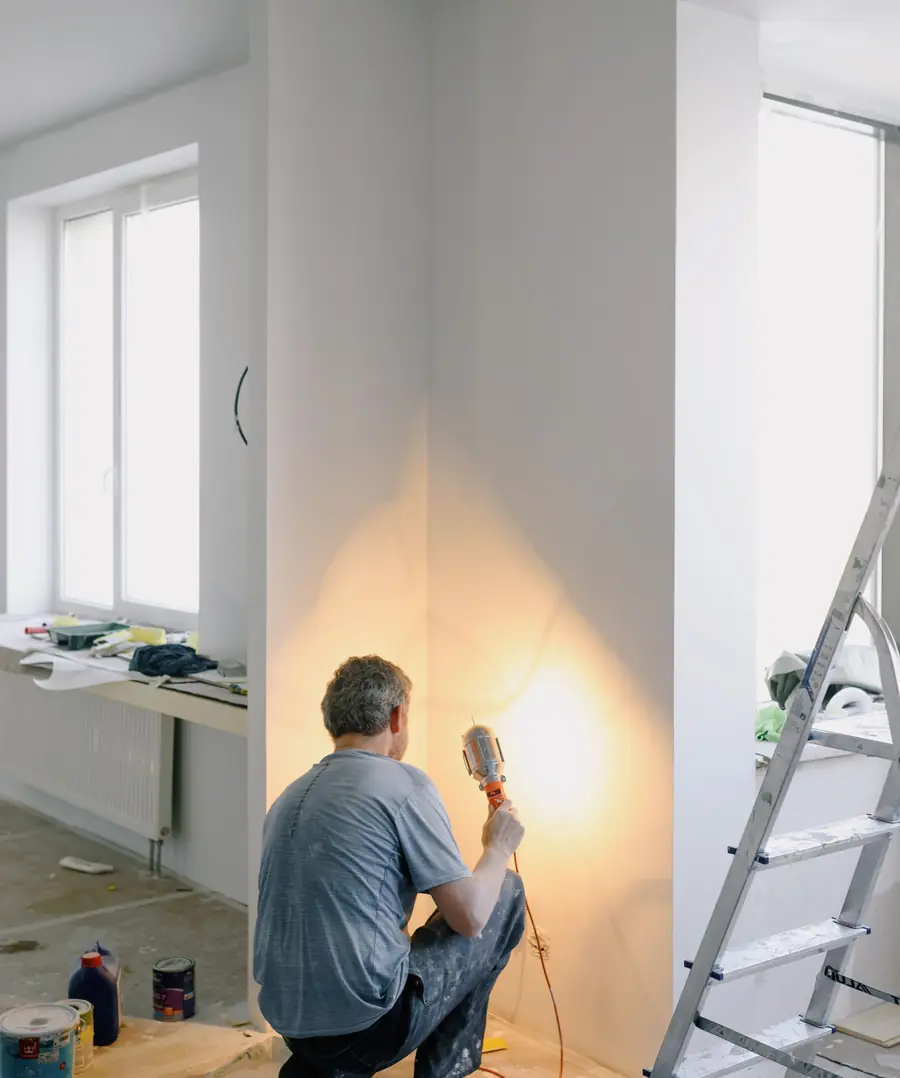 Ein Elektriker kniet mit einer Arbeitsleuchte an einer frisch verputzten Wand in einer leeren Wohnung.