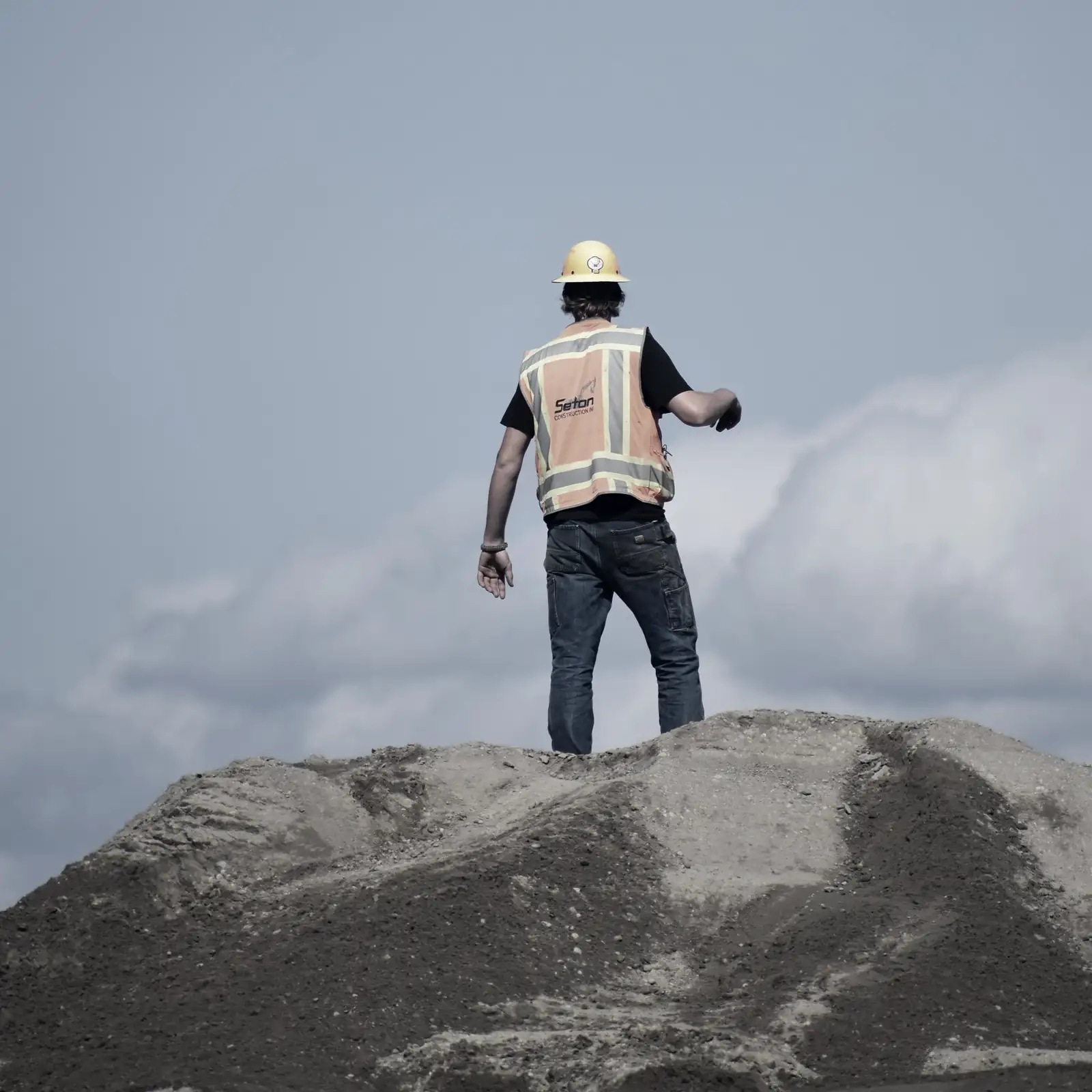 Ein Mitarbeiter in Warnweste steht von hinten auf einem Erdhaufen und zeigt über die Baustelle