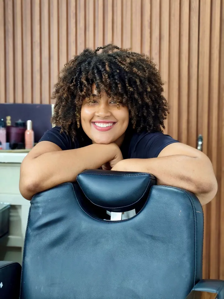 Teresa Kolar mit dunklen Locken, lachend, die Arme auf der Lehne eines Friseurstuhls.