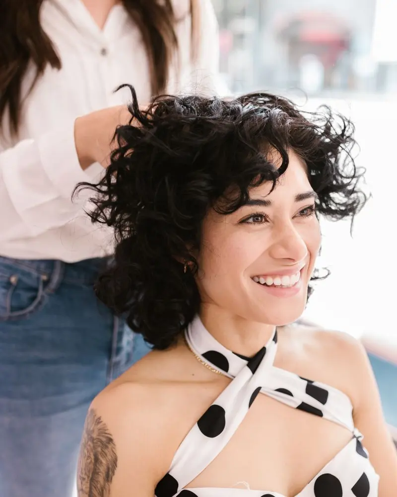 Lachende Frau mit schwarzen Locken im Salon, hinter ihr die Friseurin.