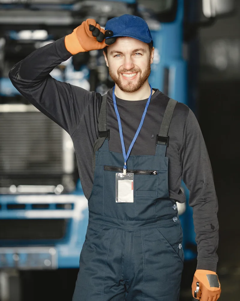 Kfz-Techniker mit Kappe und Latzhose grüßt vor einem Fahrzeug in der Werkstatt.